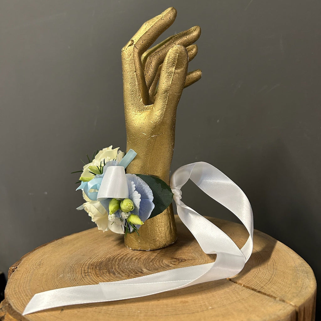 A wrist corsage with white and light blue flowers, green buds, and a white ribbon, displayed on a gold hand mannequin atop a rustic wooden stump against a dark gray background.
