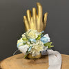 A wrist corsage with white and cream flowers, light blue and white ribbons, and green foliage, displayed on a gold hand mannequin atop a rustic wood slice against a dark gray background.