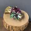 A purple and pink artificial flower wristlet or corsage with green leaves, displayed on a gold-toned decorative stand atop a rustic wood slice against a gray wall.