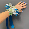 A wrist corsage with white and light green artificial flowers, white and blue ribbons, and green leaves, worn on a person's wrist with their hand flat against a gray background.