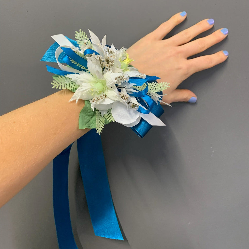 A wrist corsage with white and light green artificial flowers, white and blue ribbons, and green leaves, worn on a person's wrist with their hand flat against a gray background.