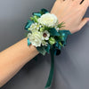 A wrist corsage featuring white roses, small white flowers, green foliage, and a dark teal ribbon bow, shown worn on a person's wrist against a plain gray background.
