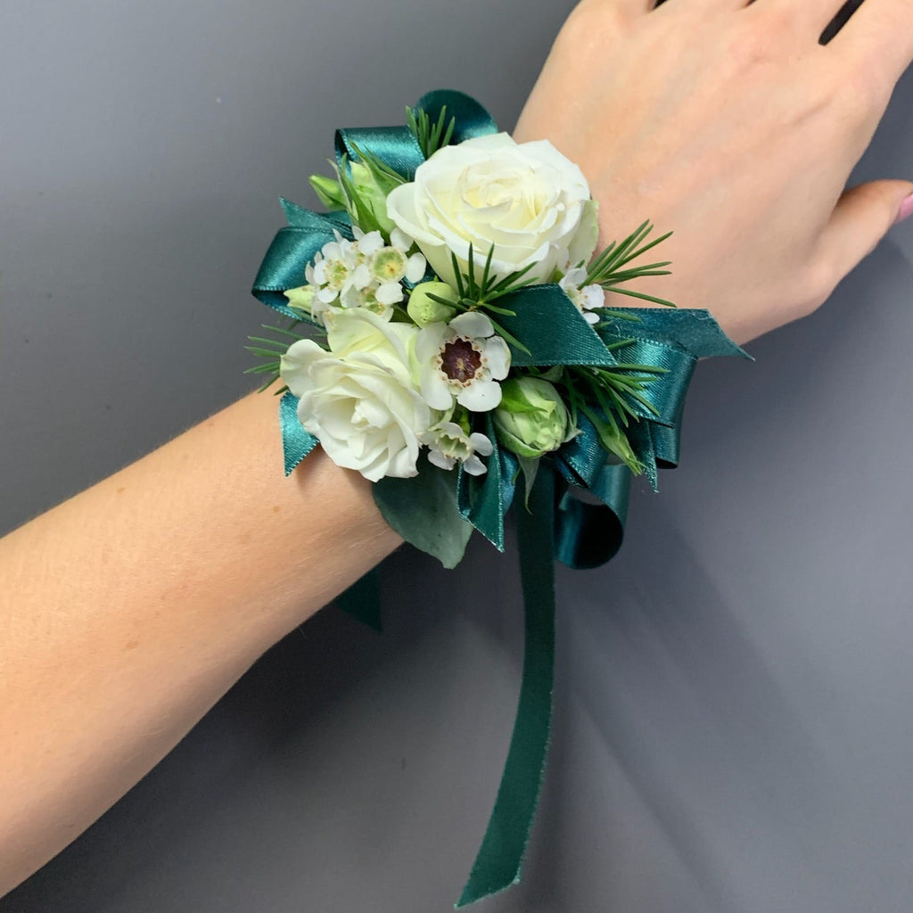 A wrist corsage featuring white roses, small white flowers, green foliage, and a dark teal ribbon bow, shown worn on a person's wrist against a plain gray background.