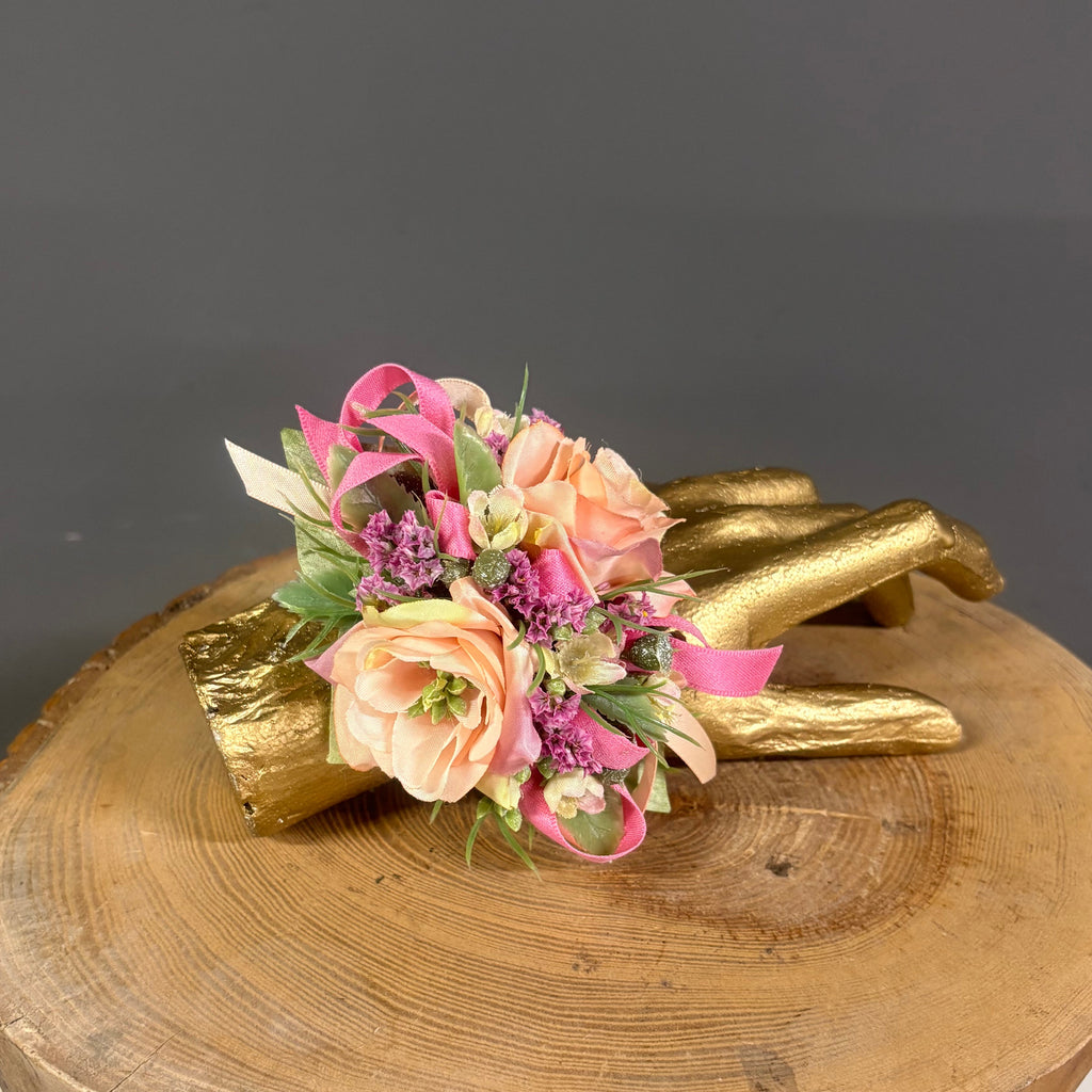 A small floral wristlet or boutonniere arrangement with peach roses, pink blossoms, purple accents, and green leaves, displayed on a gold branch over a round wood slice stand against a dark gray background.